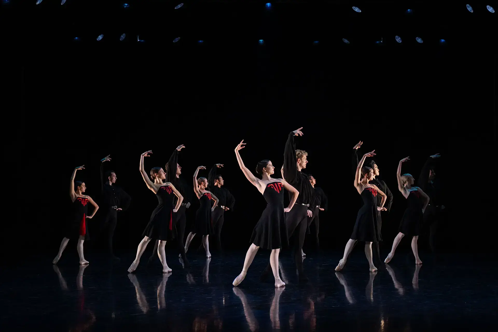 Six pair of dancers, each wearing black, the women partners with red accents on their dresses, stand posing with one hand on their hips, the other arm raise above their head