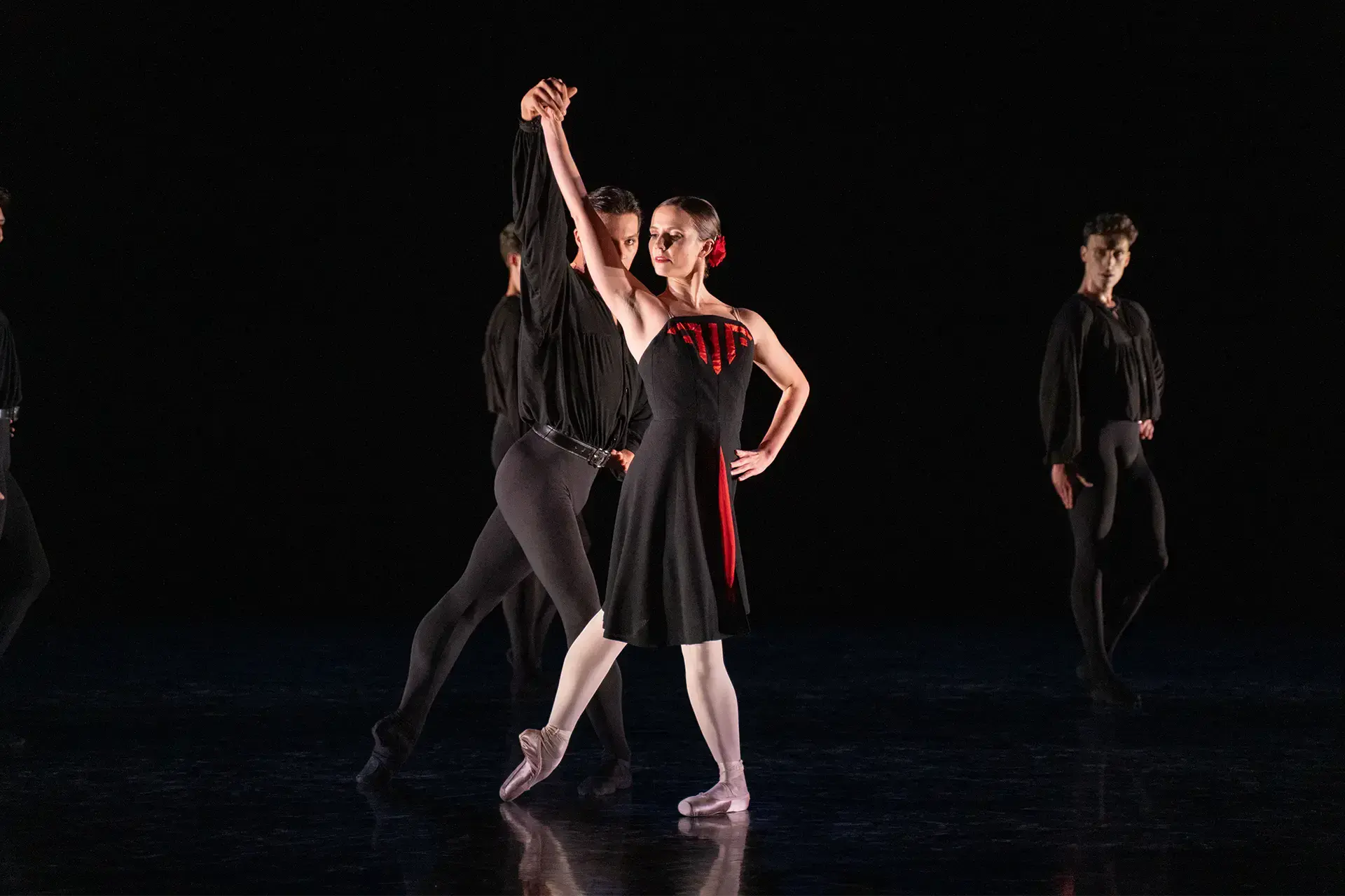 Dancer in white tights and a black off-the-shoulder dress with red accents stands against a black backdrop with her right arm raised