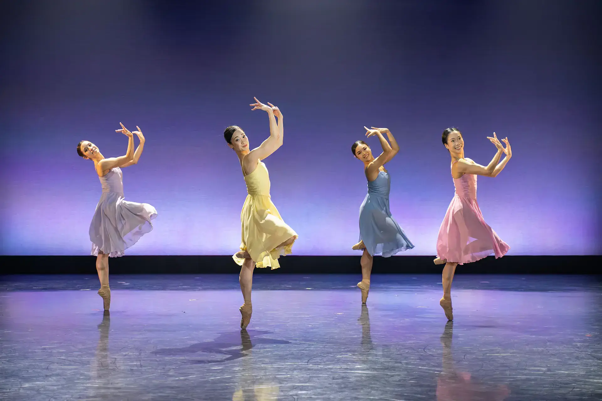 Four female dancers, one each in lilac, yellow, blue and pink, all on one foot en pointe with their arms in front of their face