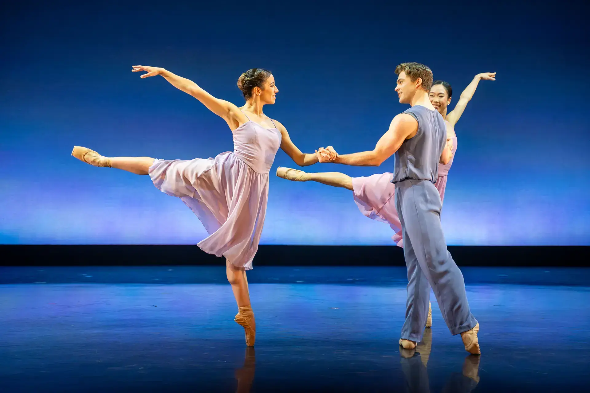 Dancers in pastel colours against a blue backdrop looking at one another charmingly