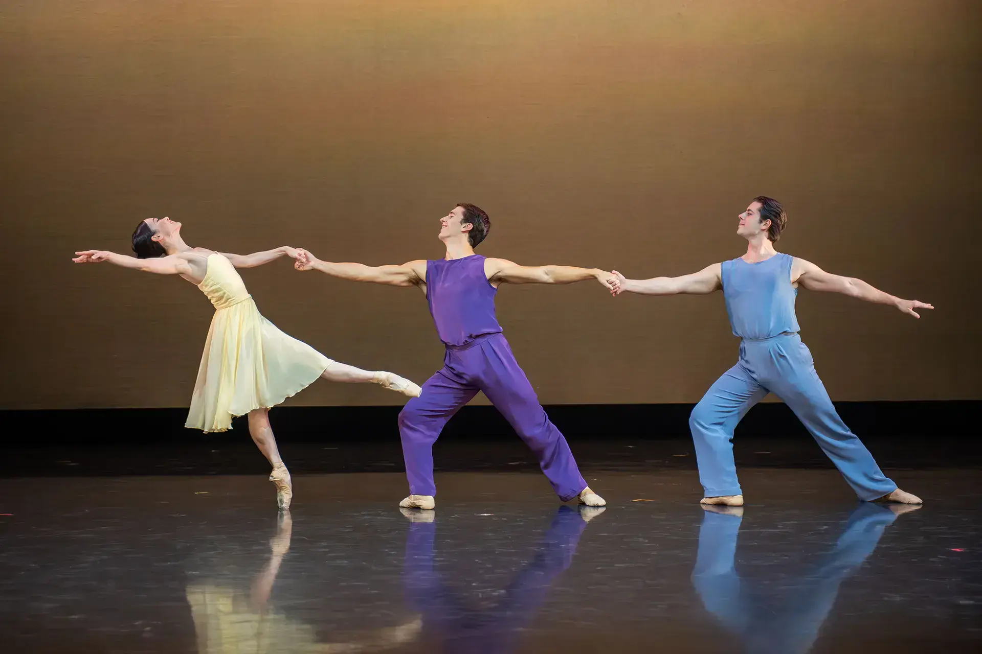 Three dancers, a woman in yellow leans back, she leans back on one foot held up by a man in purple holding her hand, he's held on to another man in blue