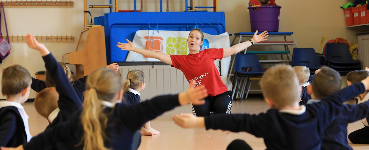 school children sat on floor with arms stretched out as teacher demonstrates in front of them