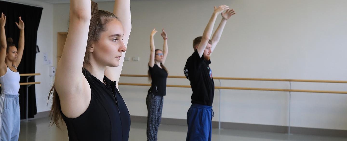 Students in a dance studio, stood still with arms raised above their heads