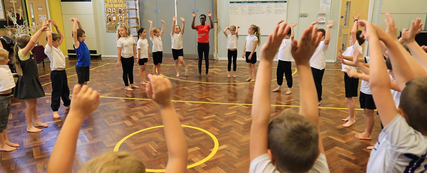 A hall full of school children standing in a circle with their arms raised in the air