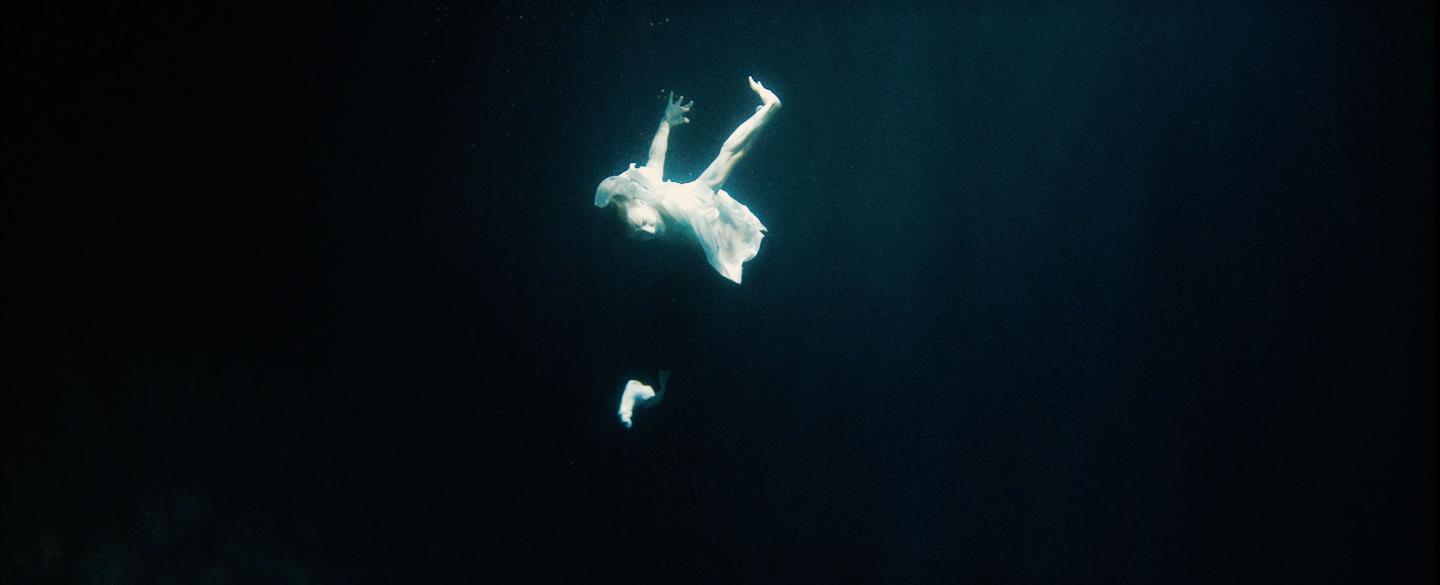 A man in a white shirt and dark trousers shapes his body like a swan whilst underwater