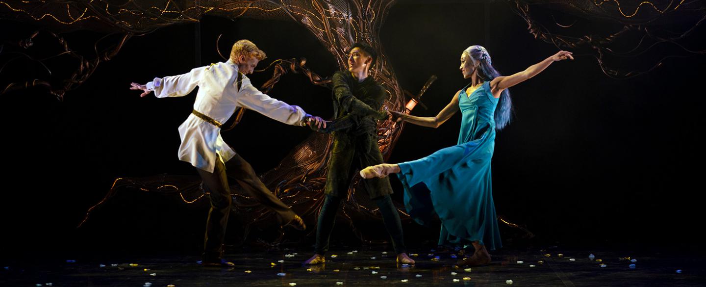 Three dancers perform in front of a large lit tree. Two dance around the other stood in the centre, holding their hands.