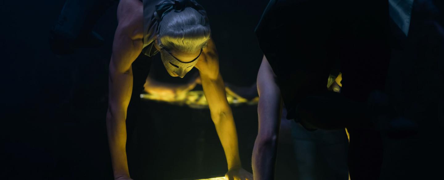 Two masked dancers look down at an illuminated chess board