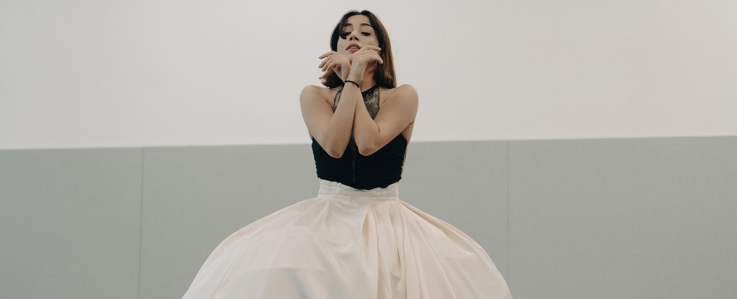 Dancer wearing a voluminous white skirt holding her forearms crossed in front of her face.