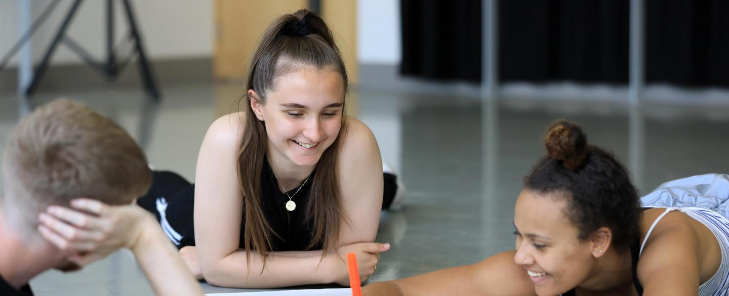 Two girls and one boy laid down on a dance studio floor. The girl on the right hand side is writing on a white piece of paper on the floor with an orange pen.
