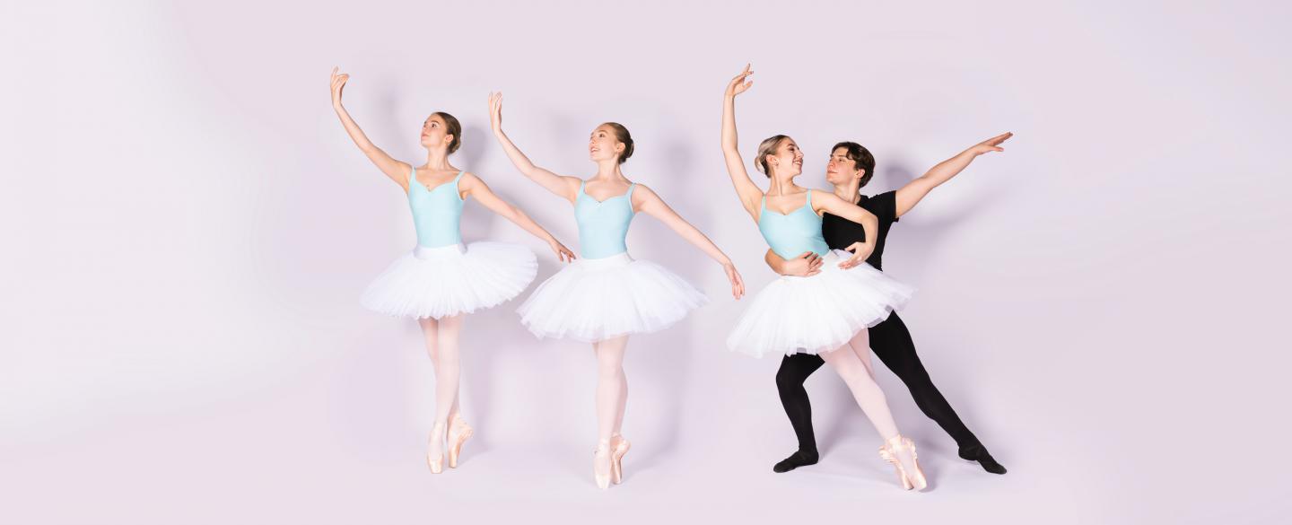 Three female dancers in white tutus stood on pointe. A male dancer holds one dancer sideways