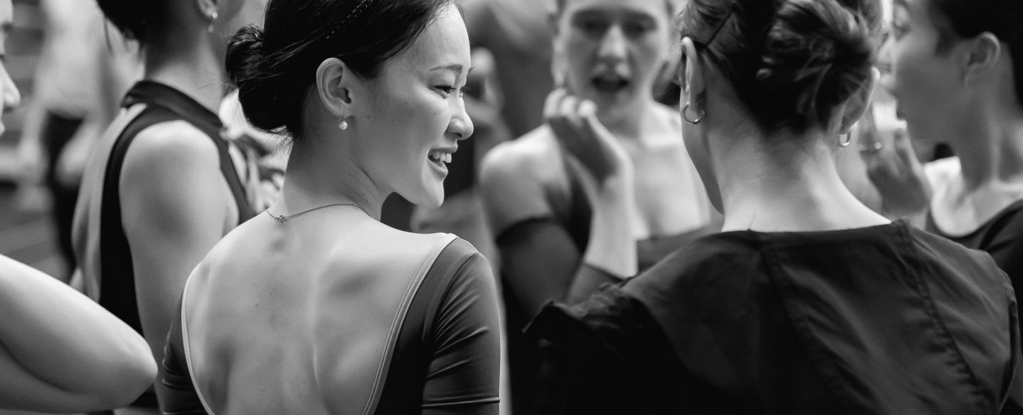 Black and white photo of a dancer in rehearsal standing in a group speaking with the person next to her