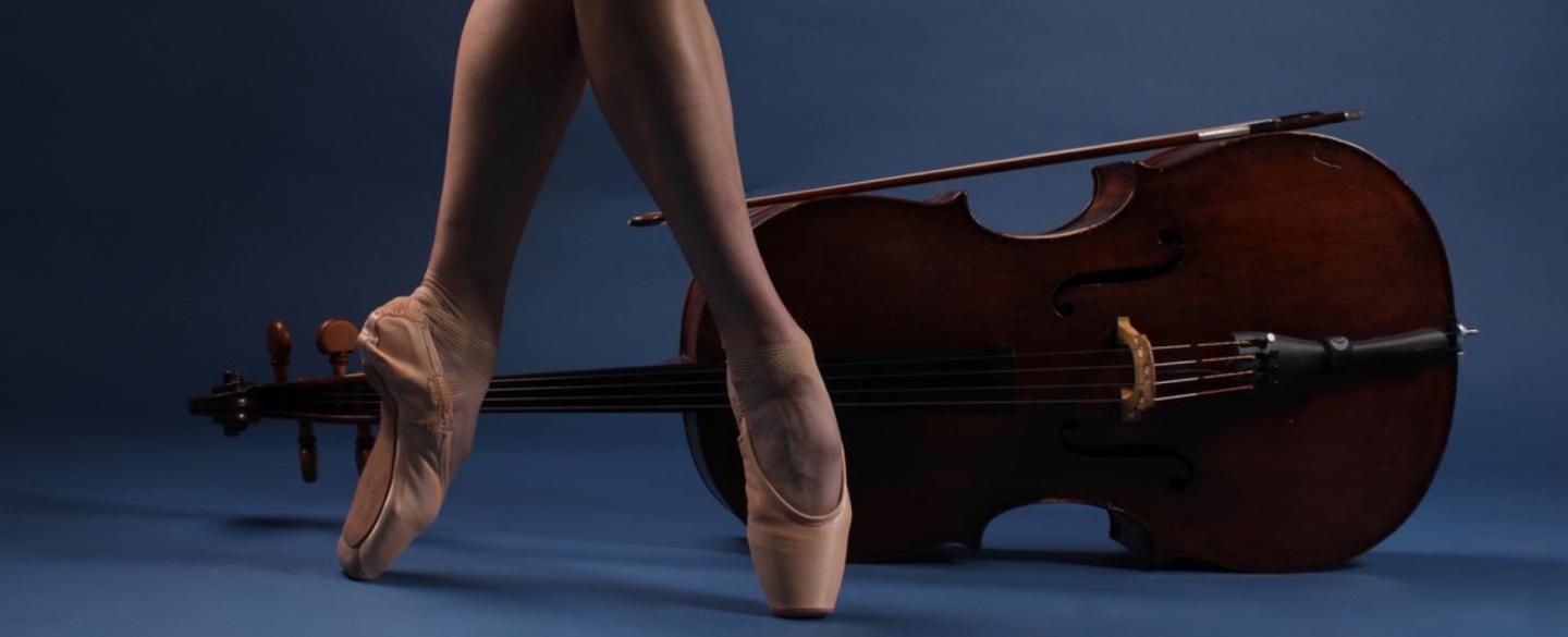 Photo of the bare lower legs wearing pointe shoes in front of a viola places on its side all against a blue background