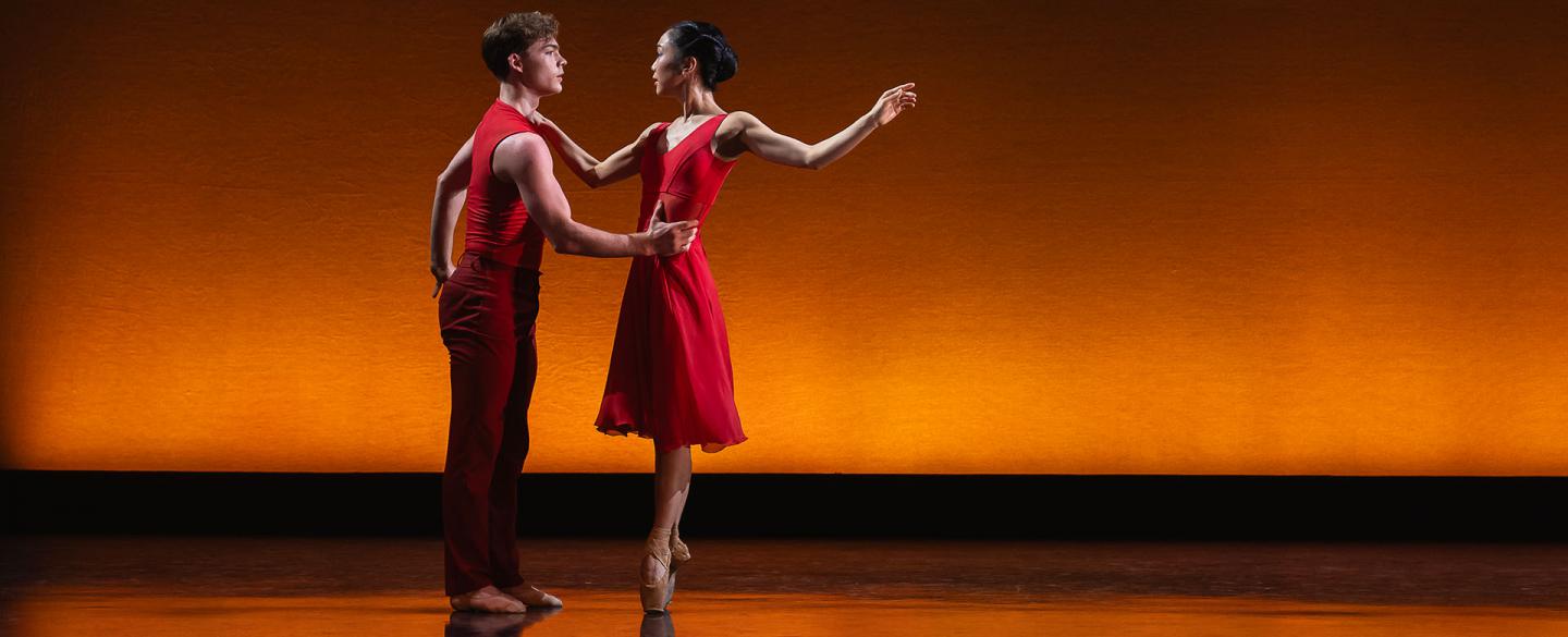 Two dancers wearing crimson, one holds the others waist and that person is en pointe, one hand on the other's shoulder