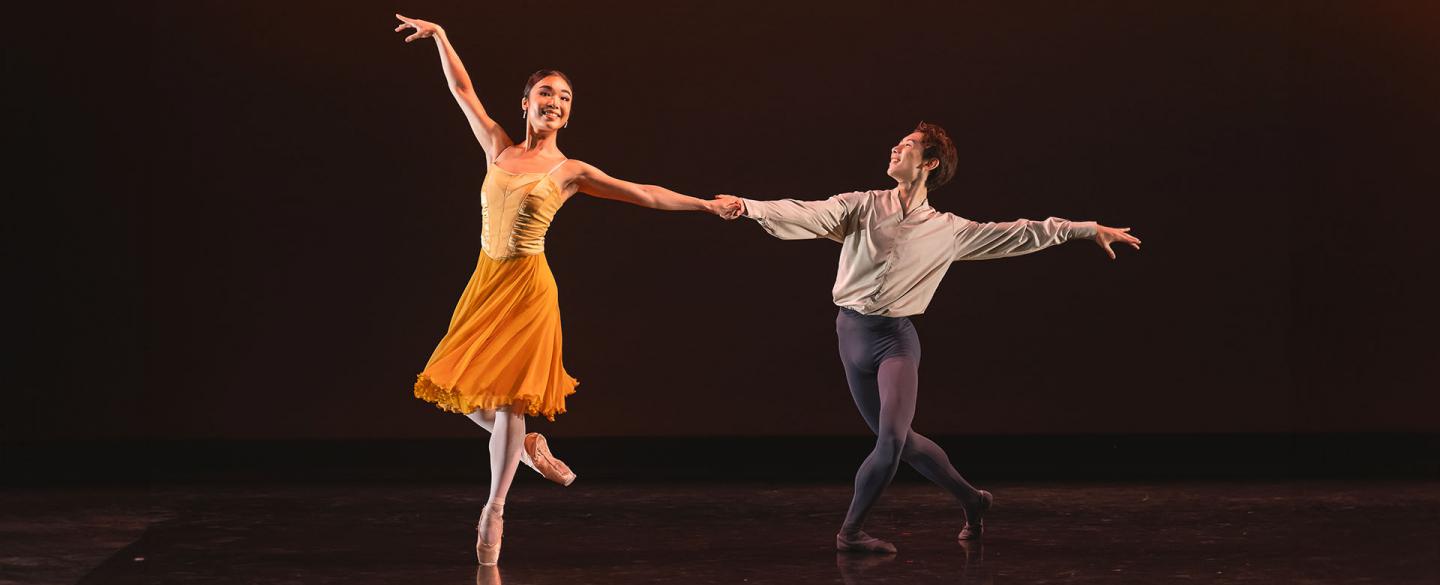 Female dancer in a orange skirt smiling as she stands on one foot en point, one leg bent at the knee behind her and an arm raised above her head while the other is held by a male dancer in white and grey