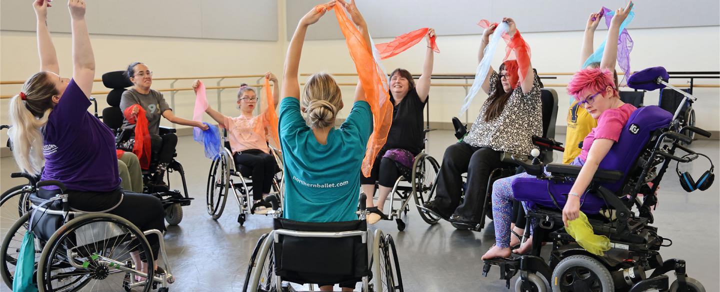 Group of dancers in wheelchairs rehearse in studio reach high holding colourful ribbons and fabric.