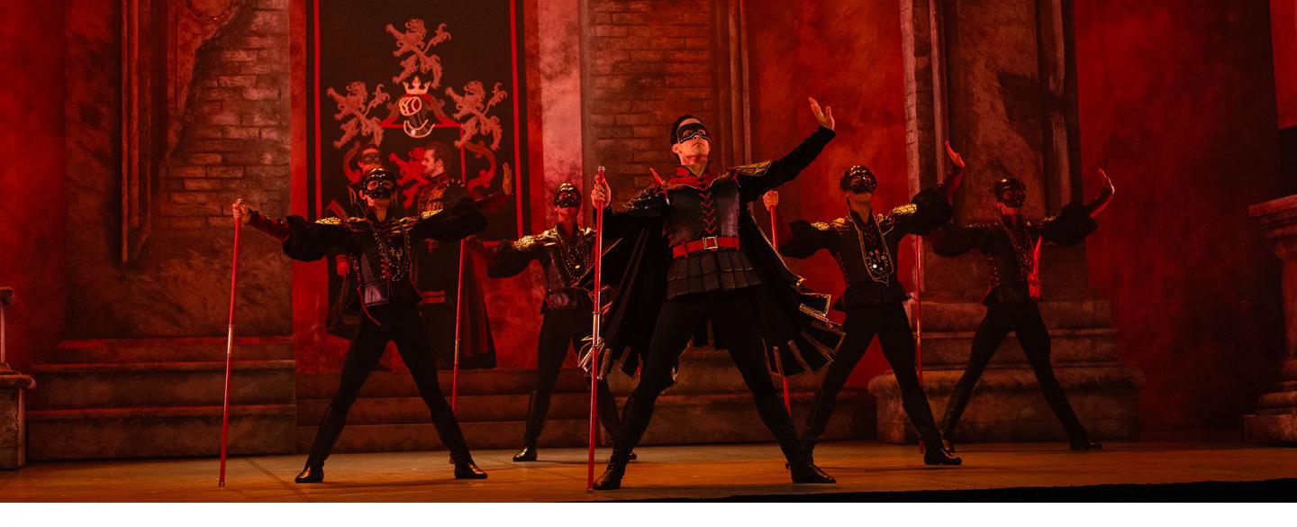 I red lighting, a group of men stand wearing masks and carrying a red staff