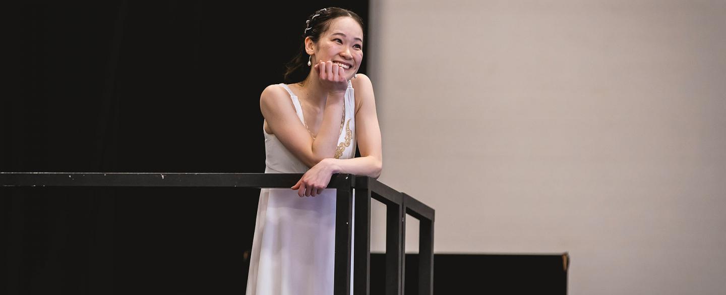 A dancer on a balcony dresses all in white smiles while resting a hand on her chin