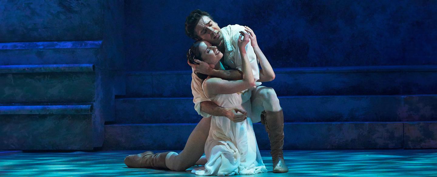 Romeo embraces Juliet as then kneel on the floor, bathed in a cool blue light