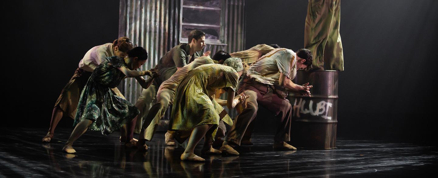A group are each leaning low, as if burdened, another stands on a metal drum shouting at them