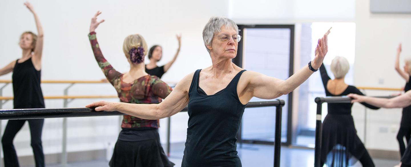 Class of dancers at the barre, one hand holding on for balance, the other arm held out, palm up forearm raised, knees slightly bent