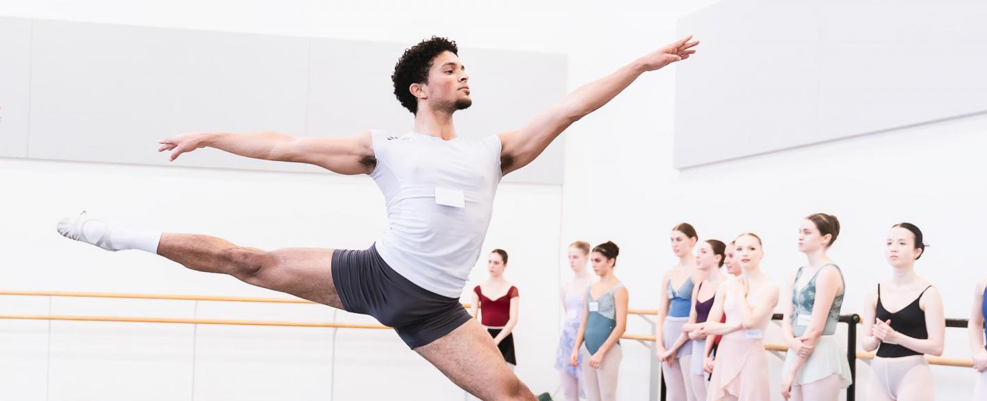 A student in a white t-shirt and black shorts leaping across a ballet studio with other students watching