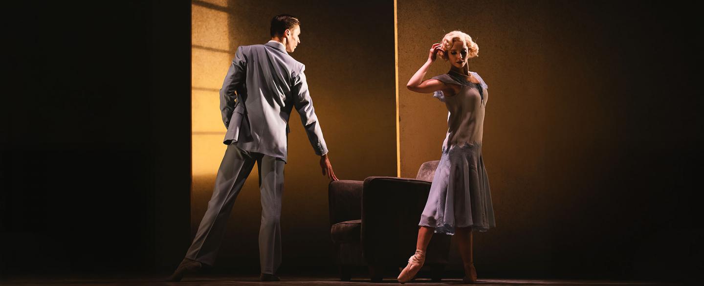 Dancers on stage in 1920 clothing, her a blue flapper dress, him is a sharp grey suit