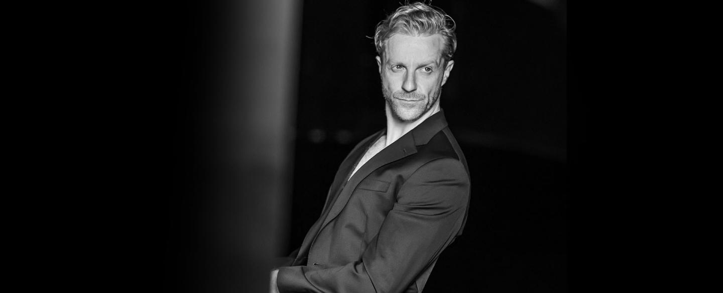 Black and white photo of blond man wearing a jacket sits leaning back looking over his shoulder.