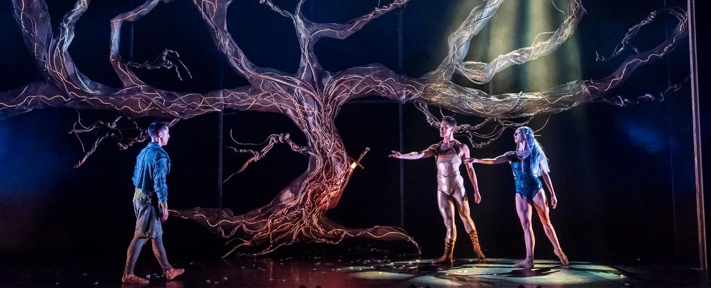 Under a leafless tree, Merlin approaches the beckoning Lady of the Lake wearing blue and Helios in golden reflective clothes.