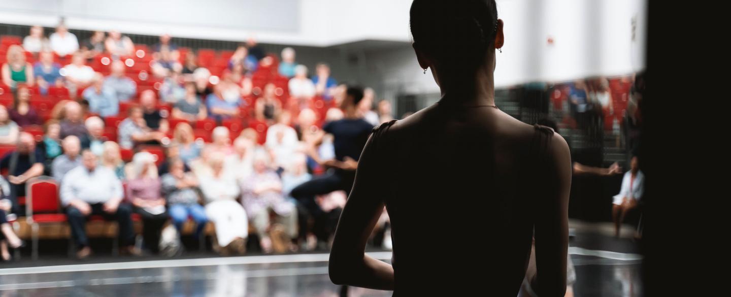 A silhouette of a dancer stood with their back to the camera while taking ballet class. On the background, a group of spectators watch from red chairs.