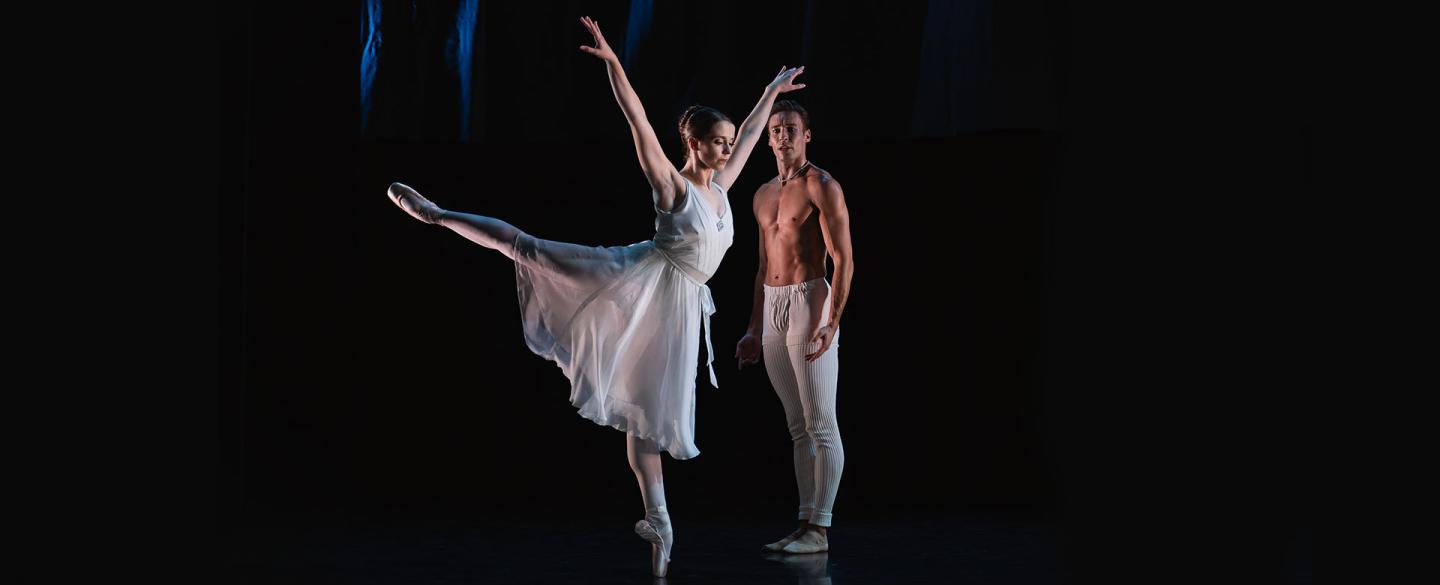 Woman in a white dress, en pointe on one foot, the other leg arabesque, both arms raise, man in white trousers stood behind her, watching.