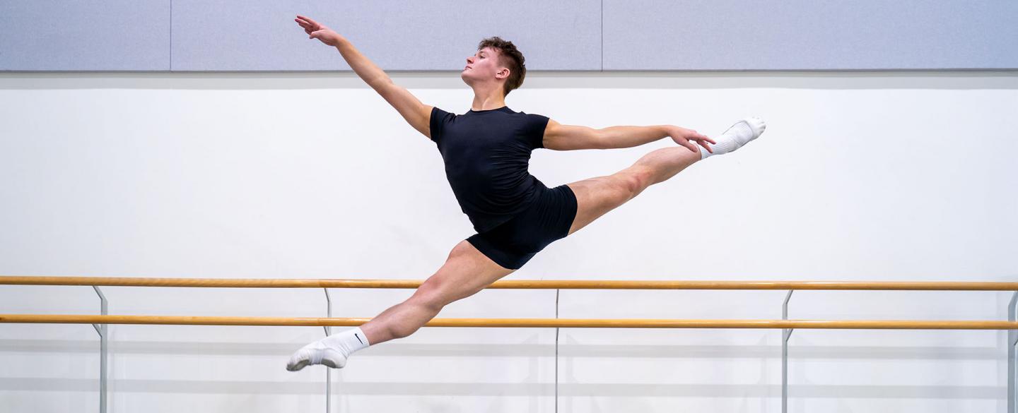 Dancer leaping across the frame in a black T-shirt and shorts, looking powerful as he stretches is legs and arms in front and behind.