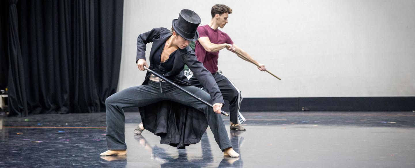 Rehearsing the role of Anne Lister, a female dancer wearing a long black coat and top hat stands, knees bent almost in a crouch, she looks down hiding her full face
