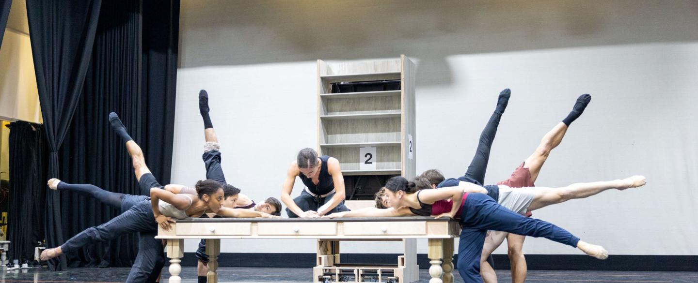 A dancer in black sits on a wooden table, leaning over whilst writing with a feather quill. Multiple dancers surround the table, each with one leg elevated behind them, reaching towards the quill.