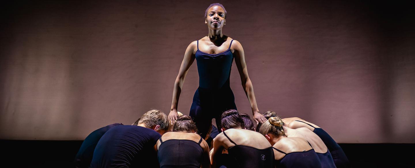 Against a dim backdrop a lone girl stands surrounded by people kneeling at her feet
