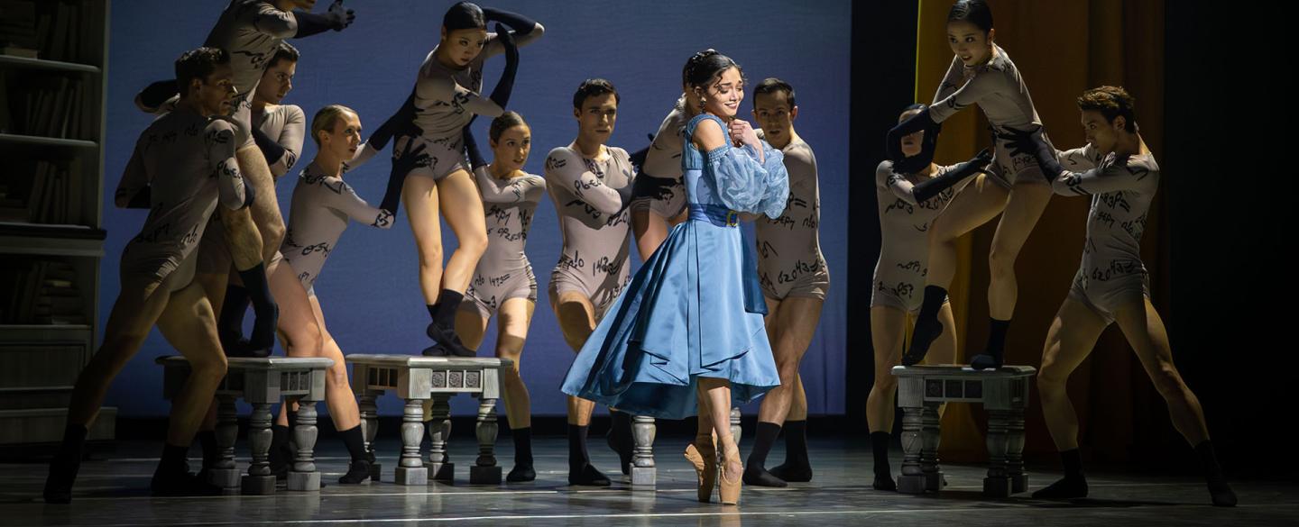 Woman in a blue dress smiles, smiling holding her hands to her chest, behind her dancers representing Anne Lister's beguiling words