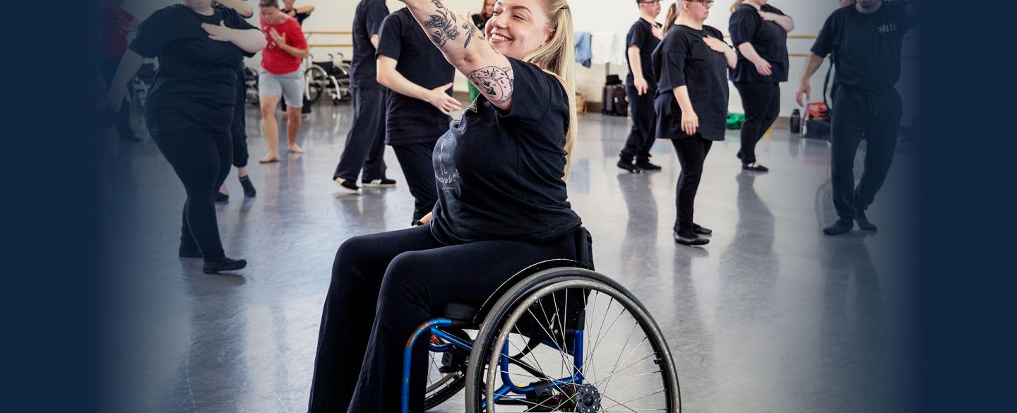 Woman in wheelchair smiling and raising her arm expressively. Young people in the background moving in unison