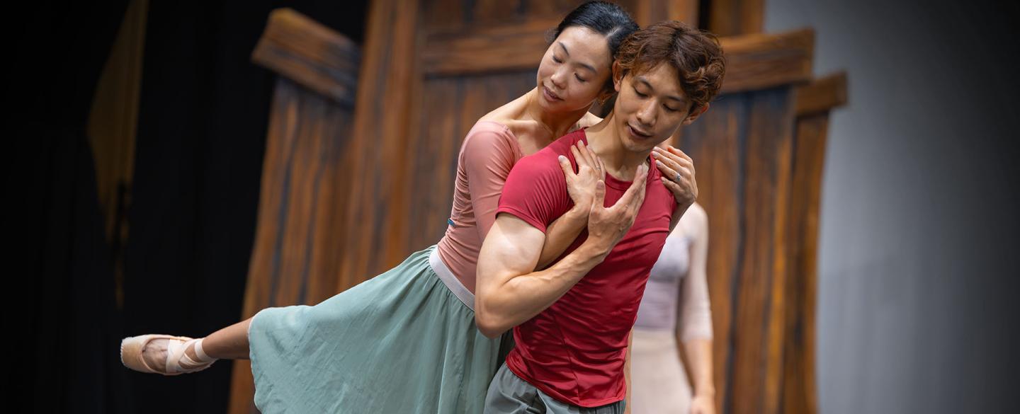 Dancer in a red t-shirt move forward, another holds him from behind and is lifted as she hooks her arms under his and holds his shoulders
