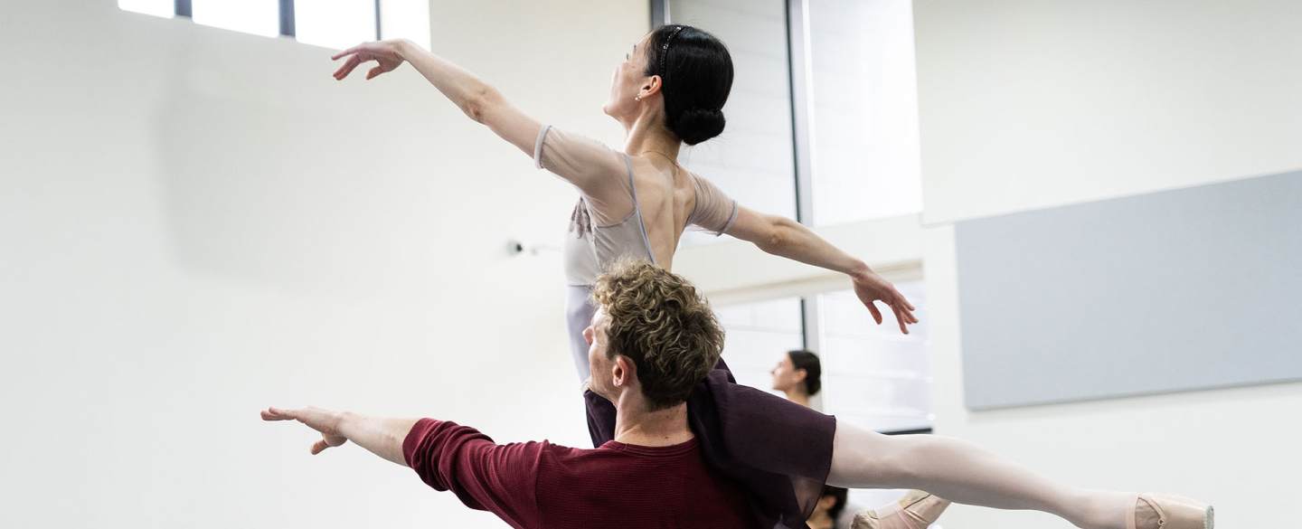 Dancer in a deep red top lifts another on his shoulder, the lifted person gracefully arches her back up, extends a hand in front of her and one leg behind