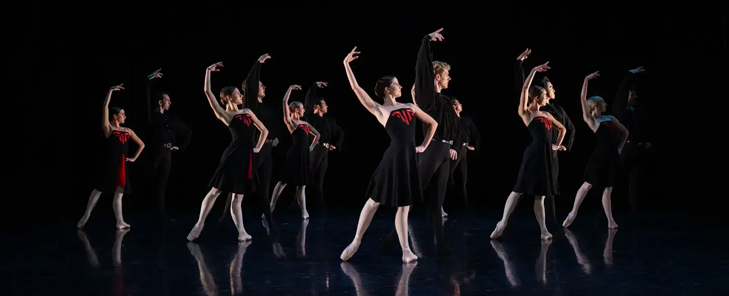 Six pair of dancers, each wearing black, the women partners with red accents on their dresses, stand posing with one hand on their hips, the other arm raise above their head