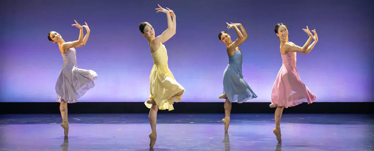 Four female dancers, one each in lilac, yellow, blue and pink, all on one foot en pointe with their arms in front of their face