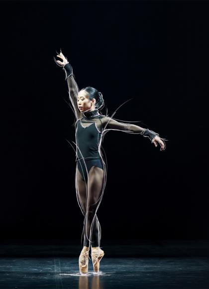 Dancer in a black leotard stands en pointe against a black background with one arm extended above her and one to her side.
