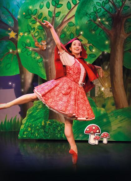 Dancer in red with a red hood and red pointe shoes stands arabesque smiling as she walks through a forest