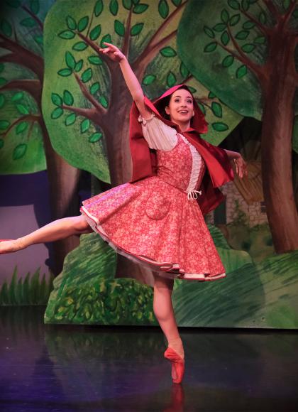 Dancer in red with a red hood and red pointe shoes stands arabesque smiling as she walks through a forest