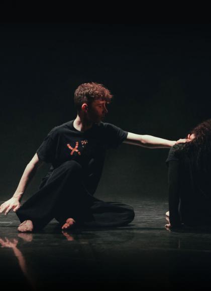 Dancers on the dark floor , lit sparingly from above, one reaching out to the other who has their back to us and head down