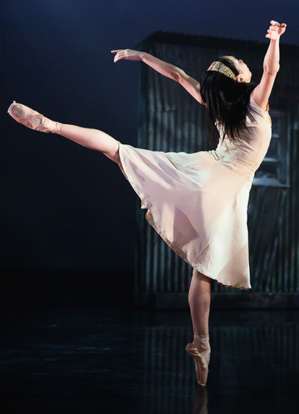 Dancer in a pale dress wearing a hair band stands on one foot en pointe, a leg straight behind her, both arms above her head