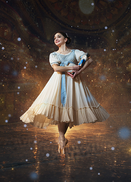 A dancer in Edwardian costume hold a Nutcracker doll in her arms, stands en pointe and smiles with childlike wonder