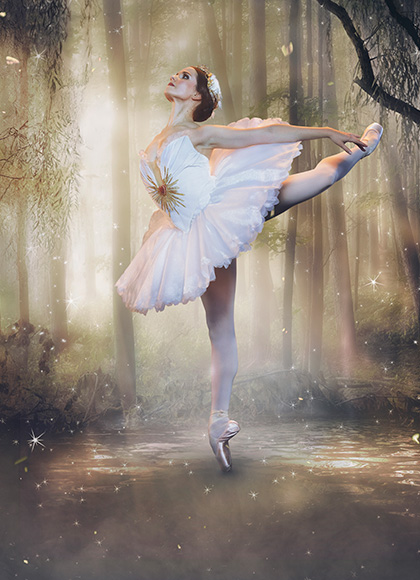 In the middle of a dark forest with soft sunlight, a dancer in a white feathery tutu poses in an attitude, with her back arched and her arms reaching to the back.