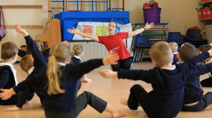 school children sat on floor with arms stretched out as teacher demonstrates in front of them