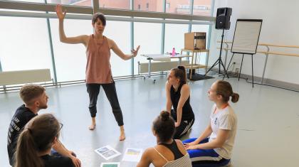 A teacher stands presenting with arms whilst five people sit on the floor watching