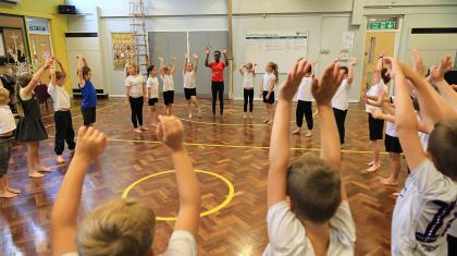 A hall full of school children standing in a circle with their arms raised in the air